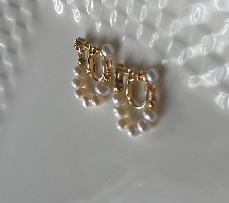 May include: A pair of gold-toned clip-on earrings with a pearl design. The earrings are made of small, white pearls arranged in a circular shape.