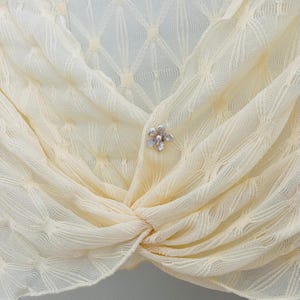 May include: Cream-colored, open-weave shawl with a delicate floral brooch. The shawl is knotted at the front and features a subtle diamond pattern. The brooch is silver-toned with pearl-like accents, adding a touch of elegance to the soft fabric.