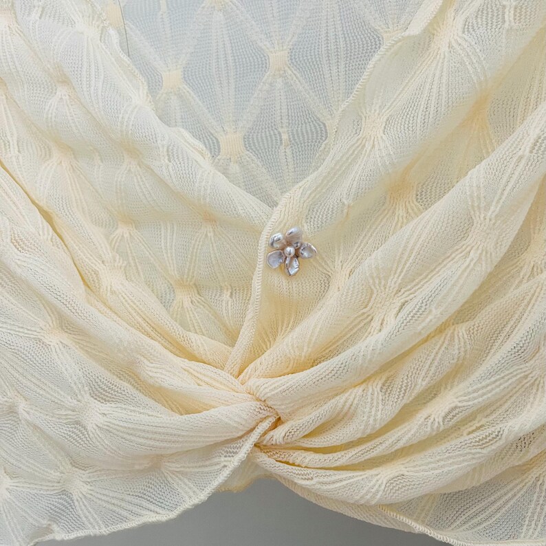 May include: Cream-colored, open-weave shawl with a delicate floral brooch. The shawl is knotted at the front and features a subtle diamond pattern. The brooch is silver-toned with pearl-like accents, adding a touch of elegance to the soft fabric.