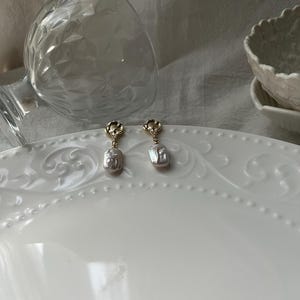 Puede incluir: Un par de pendientes de oro con una perla colgante. Los pendientes tienen un dise&ntilde;o floral en la parte superior.