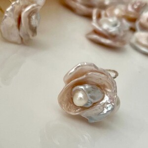 May include: A delicate pearl flower charm with a white pearl in the center. The flower is made of iridescent pink and white shell and has a gold wire loop for attaching to jewelry.
