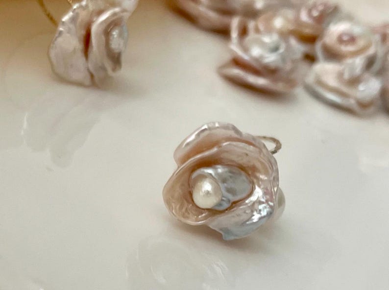 May include: A delicate pearl flower charm with a white pearl in the center. The flower is made of iridescent pink and white shell and has a gold wire loop for attaching to jewelry.