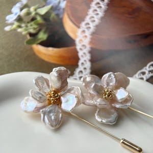 Puede incluir: Dos broches de flores de perlas blancas con detalles dorados. Los broches est&aacute;n sobre una superficie blanca y tienen un alfiler en la parte posterior.