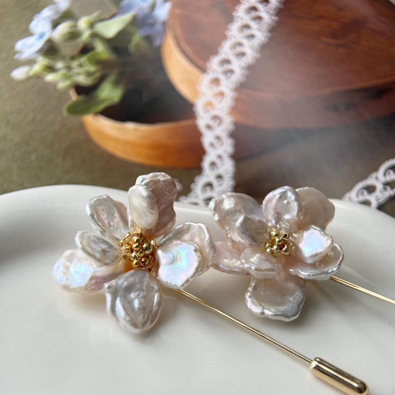 Puede incluir: Dos broches de flores de perlas blancas con detalles dorados. Los broches est&aacute;n sobre una superficie blanca y tienen un alfiler en la parte posterior.