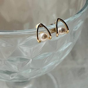 K&ouml;nnte beinhalten: Ein Paar goldene Ohrringe mit einem einfachen, geschwungenen Design und einem kleinen wei&szlig;en Perlenakzent an jedem Ohrring.