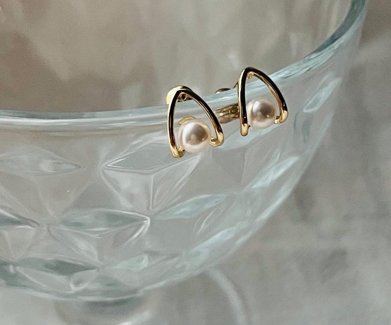 K&ouml;nnte beinhalten: Ein Paar goldene Ohrringe mit einem einfachen, geschwungenen Design und einem kleinen wei&szlig;en Perlenakzent an jedem Ohrring.