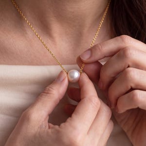 Puede incluir: Un colgante de perla blanca redonda individual suspendido de una delicada cadena de oro. La perla presenta una superficie lisa y brillante, centrada en el collar de eslabones metálicos finos que descansa sobre una prenda de tela clara.