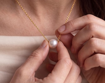May include: A single round white pearl pendant suspended on a delicate gold chain. The pearl features a smooth, lustrous surface and is centered on the thin metallic link necklace, which rests against a light-colored fabric garment.
