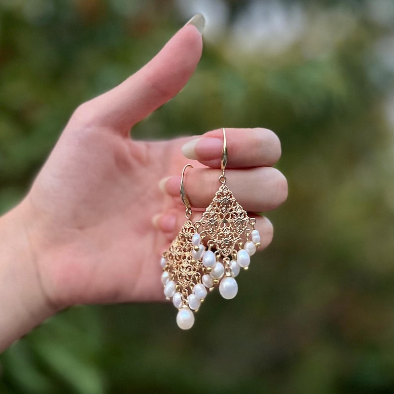Peut inclure: Boucles d'oreilles pendantes dor&eacute;es avec un motif en filigrane m&eacute;tallique ajour&eacute; en forme de losange. Chaque boucle est orn&eacute;e de plusieurs petites perles d'eau douce blanches en forme de goutte suspendues au bord inf&eacute;rieur.