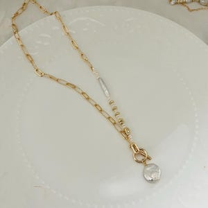 May include: Gold-coloured chain necklace with a mix of oval and rectangular links. The necklace features a strand of small pearls, a silver-coloured bar, and a large, irregular-shaped pearl pendant. A gold clasp secures the pendant.