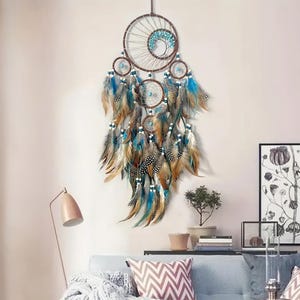 K&ouml;nnte beinhalten: Ein gro&szlig;er Traumf&auml;nger mit braunen und blauen Perlen und Federn. Der Traumf&auml;nger hat ein kreisf&ouml;rmiges Design mit einem netzartigen Muster und h&auml;ngt an einer wei&szlig;en Wand.