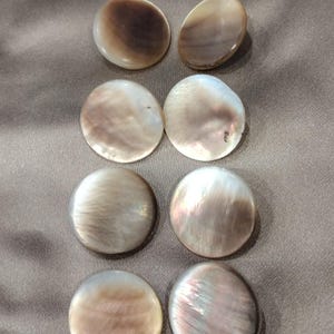 May include: Eight circular buttons arranged in two columns. Each button features a smooth, iridescent surface with varying shades of brown, cream, and grey, displaying a natural pearlescent sheen and unique swirling patterns across the round faces.