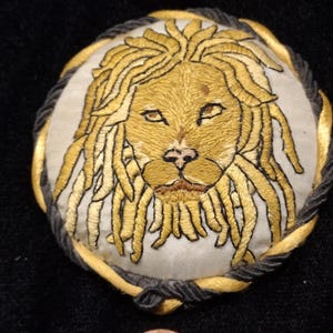 Puede incluir: Parche bordado con la cabeza de un león con hilo dorado y beige sobre un fondo blanco. La melena del león está detallada con hebras largas y fluidas. El parche está enmarcado por un borde de cuerda trenzada dorada y negra. Una moneda está en primer plano para la comparación de tamaños.
