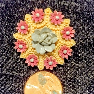 Könnte beinhalten: Dekorative Blumenbrosche mit einem zentralen grauen Sukkulenten-Design, umgeben von rosa Blüten mit weißen Zentren, auf cremefarbenem Grund. Ein Cent-Stück ist als Größenreferenz enthalten.