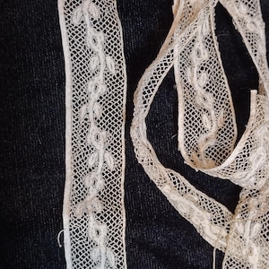May include: A long strip of off-white lace trim featuring a delicate, repeating vine and leaf pattern woven into a fine mesh background. The lace has straight, finished edges and a soft, textured appearance with a consistent, narrow width throughout.