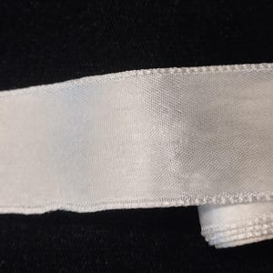 May include: A continuous length of white satin ribbon with a smooth, reflective surface and finished edges. The ribbon is partially coiled at one end, showing a consistent width and texture throughout its length, suitable for various craft projects.