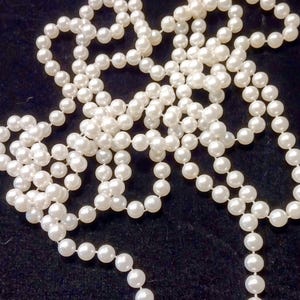 May include: A long strand of white, spherical faux pearls arranged in a loose, tangled pile. The individual beads are uniform in size and have a smooth, reflective surface, creating a continuous chain of white circular shapes against a dark background.