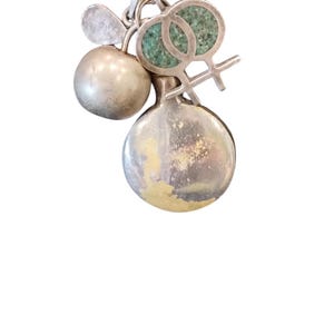 May include: Silver pendant necklace with multiple charms. The charms include a sphere, a bottle-shaped charm, and a charm with two interlocking female symbols filled with turquoise-colored material. The necklace hangs on a silver chain.