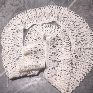 Può includere: Un colletto all'uncinetto color crema caratterizzato da un intricato motivo floreale con bordi smerlati. Il pezzo è realizzato in filo sottile, mostrando un delicato design traforato che forma una sagoma semicircolare.