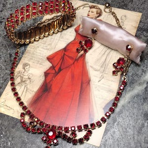 Puede incluir: Un conjunto de joyería en tono dorado que incluye una pulsera elástica y un collar con pendientes colgantes a juego. Las piezas están adornadas con numerosas piedras rojas pequeñas y facetadas y piedras centrales rojas más grandes.