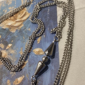 May include: A silver-toned metal chain necklace featuring multiple delicate curb chain strands. The design incorporates polished, teardrop-shaped metal pendants and small spherical beads interspersed along the length of the silver-colored chain links.
