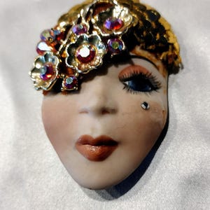 May include: A ceramic face brooch featuring a pale complexion, painted blue eyes with long lashes, and brown lips. A small rhinestone is placed below the left eye. The hair is adorned with gold-toned metal flowers set with iridescent crystals.