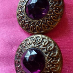 May include: Two round buttons featuring a central faceted deep purple gemstone set within an ornate antique brass-toned metal frame. The metal border displays a textured, raised lattice pattern surrounded by a decorative scalloped edge design.