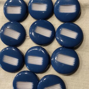 May include: A collection of eleven round, dark blue buttons arranged in a grid. Each button features a glossy finish and a central horizontal white stripe pattern that appears to have a textured, raised appearance across the surface of the button.