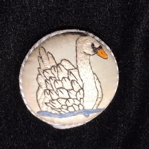 May include: A round, embroidered patch featuring a white swan with an orange beak and black accents, floating on blue water. The swan is set against a cream-colored background, with a white stitched border.