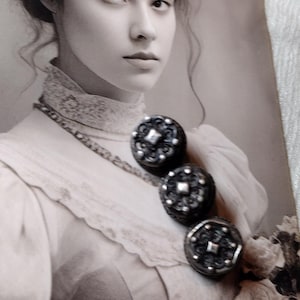 Puede incluir: Una fotografía en tono sepia de una persona con el cabello recogido que viste una blusa de encaje de cuello alto. Tres botones decorativos circulares oscuros con detalles metálicos plateados están prendidos en el pecho.