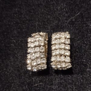 Op de afbeelding: Een paar zilverkleurige oorringen met vijf verticale rijen van heldere, gefacetteerde stenen in een rechthoekig, gebogen ontwerp. De oorbellen hebben een metallic afwerking en een gestructureerd uiterlijk met meerdere rijen.