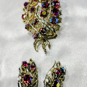 May include: A gold-tone metal jewelry set consisting of a brooch and a pair of matching earrings. Each piece features a swirling leaf-like design accented with round, multi-faceted iridescent crystals in shades of deep red, purple, and blue.