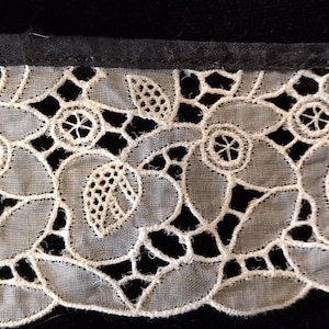 Puede incluir: Primer plano de un ribete de encaje blanco con un diseño floral. El diseño presenta flores y hojas estilizadas, con detalles calados. El encaje se coloca sobre un fondo oscuro, resaltando el delicado patrón.