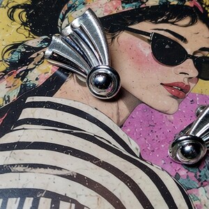 May include: A pair of silver-toned metal earrings featuring a fan-shaped design with fine vertical etched lines and a smooth, polished spherical accent at the base. The earrings have a screw-back mechanism visible on the side of the metal structure.