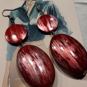 May include: A pair of drop earrings featuring a small circular disc connected to a larger oval-shaped pendant. Both components are finished in a deep red hue with a brushed, streaked texture in lighter tones and attached to silver-toned metal hooks.