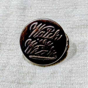 May include: A circular metal pin with a dark, oxidized finish. The surface features embossed, raised lettering in a cursive script that reads "Watch the Mess". The edges of the pin are smooth and rounded, creating a distinct contrast between the dark recessed areas and the lighter, metallic raised text.