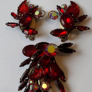 May include: A vintage jewelry set featuring a brooch and clip-on earrings. The set showcases a floral design with deep red, oval-cut rhinestones and iridescent accents. The brooch and earrings have a clip-on closure.