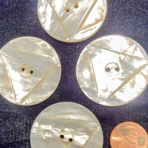 May include: Four round, pearlescent buttons with a triangle design and two holes for sewing. A copper penny is included for size comparison. The buttons are on a blue denim background.