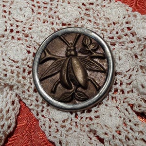 Può includere: Un bottone circolare caratterizzato da un design in rilievo color bronzo raffigurante un insetto con ali e antenne, circondato da un bordo metallico liscio color argento. L'insetto è raffigurato poggiato su piccole foglie e rami.