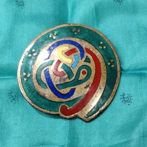 May include: A circular metal brooch featuring a central knotwork design in blue, red, and yellow enamel. The outer border is finished in green enamel with small gold-toned dot accents. The metal surface shows a weathered, aged patina throughout.