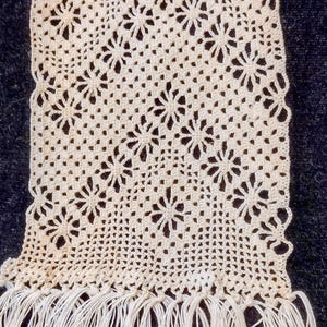 Peut inclure: Un napperon rectangulaire en crochet blanc cassé présentant un motif floral répétitif disposé en rangées diagonales. La pièce est réalisée avec un fil fin et terminée par une longue frange droite sur le bord inférieur, créant un aspect texturé.
