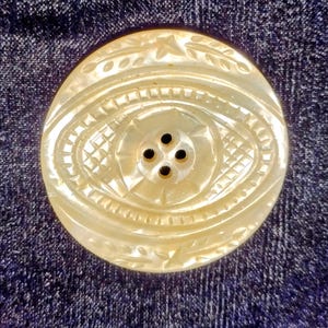 May include: A large, oval mother-of-pearl button with detailed carvings. The button has a central eye-shaped design with four sewing holes, encircled by geometric and floral patterns. It is set against a dark blue fabric.