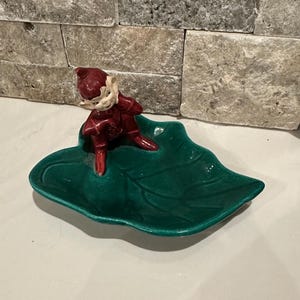 May include: A ceramic figurine of a red-clad elf on a green leaf-shaped dish. The elf has a white face. The dish is a vibrant green with a glossy finish. The figurine is in a playful pose.