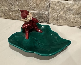 May include: A ceramic figurine of a red-clad elf on a green leaf-shaped dish. The elf has a white face. The dish is a vibrant green with a glossy finish. The figurine is in a playful pose.