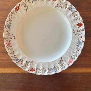Puede incluir: Un plato de cerámica blanca con una textura tejida alrededor del borde. El borde está decorado con un estampado floral con flores rojas, azules y rosas con hojas verdes. El plato está sobre una superficie de madera oscura.