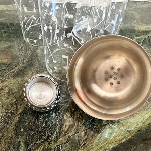 May include: A stainless steel cocktail shaker with a separate cap and a strainer lid, accompanied by clear glass tumblers featuring a white printed pattern of golf clubs and golf balls, arranged together on a surface.