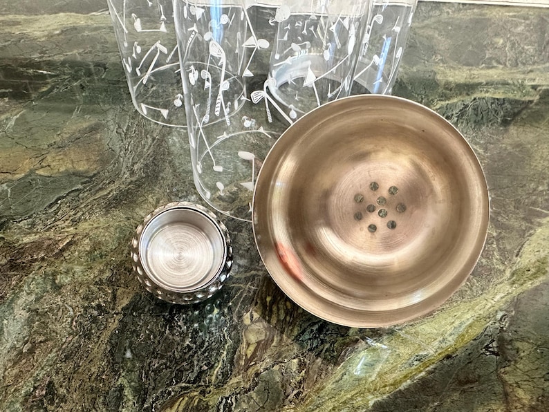 May include: A stainless steel cocktail shaker with a separate cap and a strainer lid, accompanied by clear glass tumblers featuring a white printed pattern of golf clubs and golf balls, arranged together on a surface.