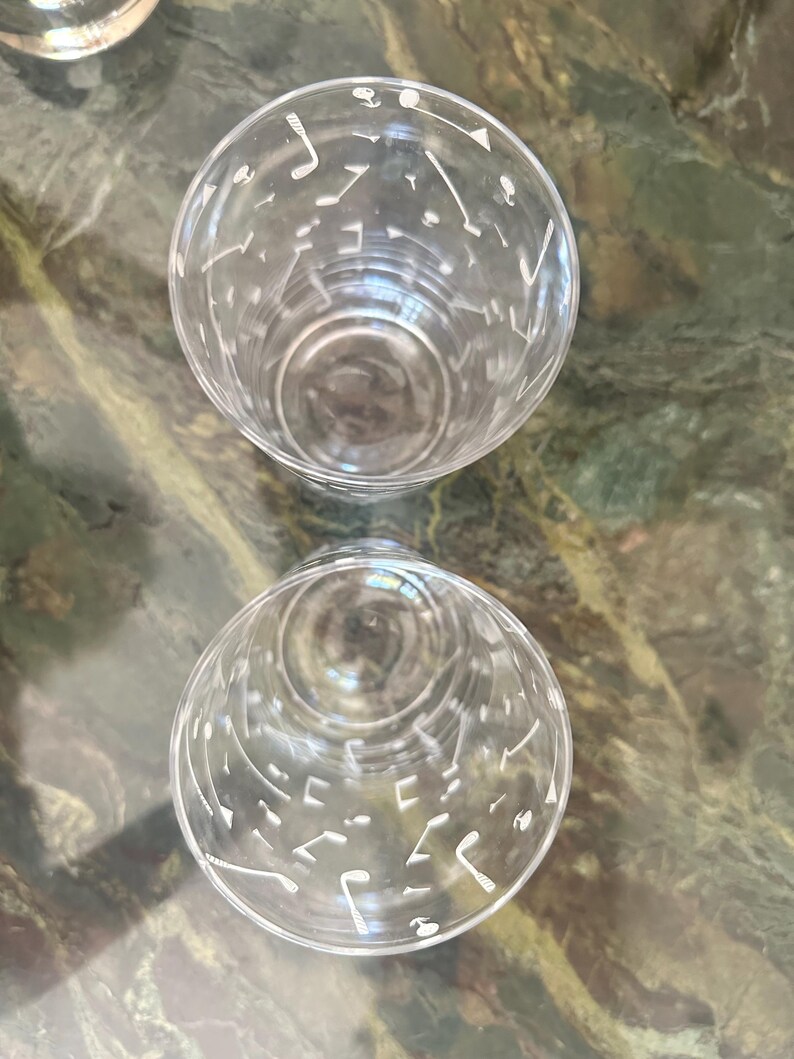 May include: Two clear glass tumblers viewed from above, each featuring a repeating white pattern of golf clubs, golf balls, and tees printed around the exterior surface of the cylindrical vessels with open circular rims.