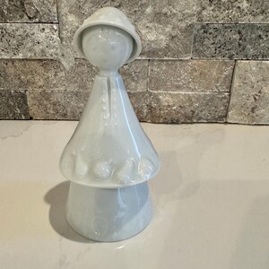 May include: A white ceramic figurine with a mushroom-shaped base and a figure wearing a hat and coat. The figurine has a smooth, glossy finish and is set against a neutral background.
