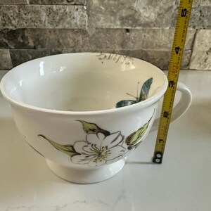 May include: White ceramic bowl with a handle, featuring a floral and butterfly design in green and blue. The bowl is approximately 4 inches in diameter and 3 inches tall. The bowl has a pedestal base.